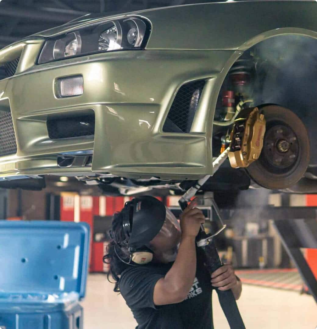 A masked man operates a pressure washer, cleaning a car tire to remove dirt and enhance its appearance