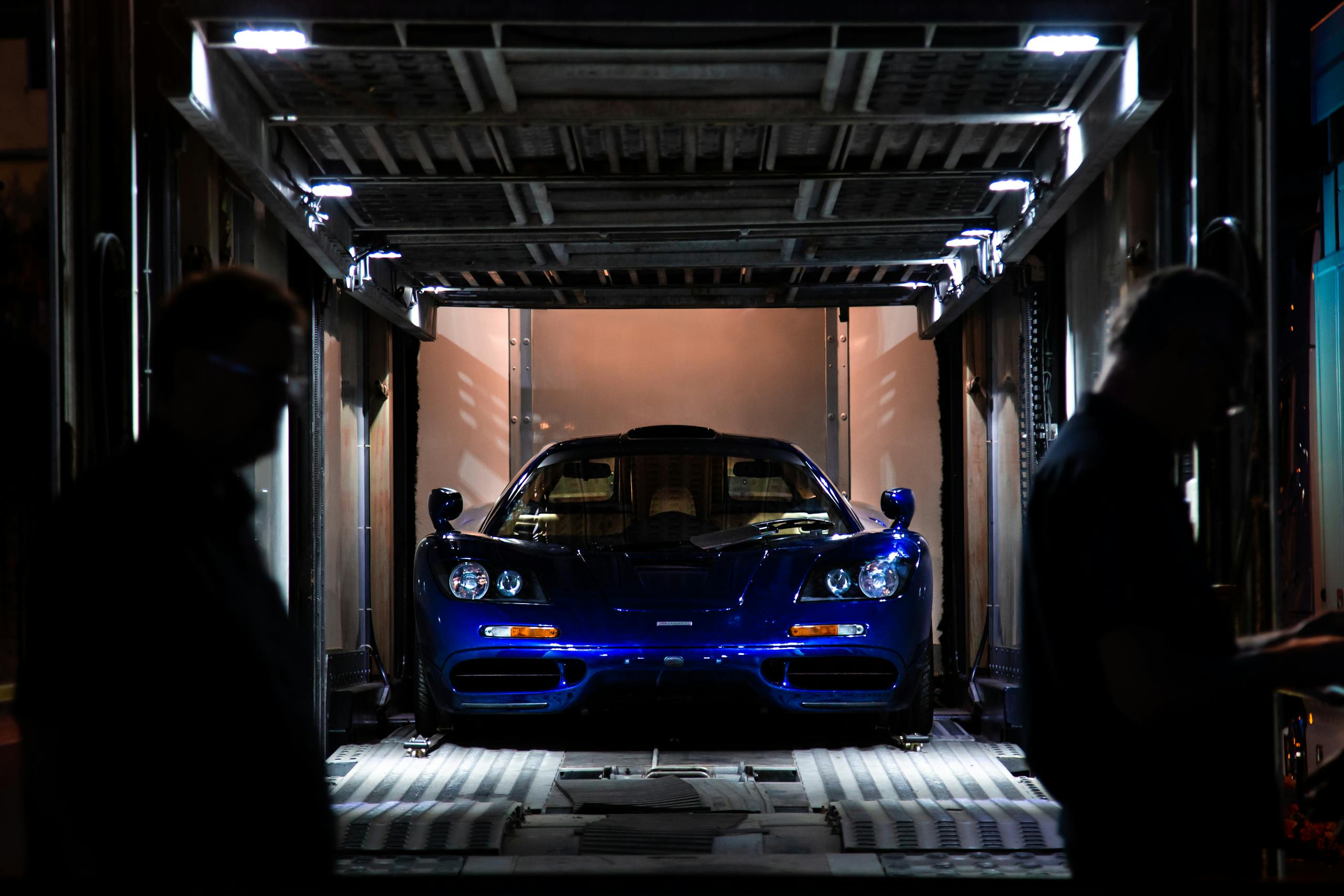 A stunning blue supercar inside a transport container, silhouetted figures in foreground.